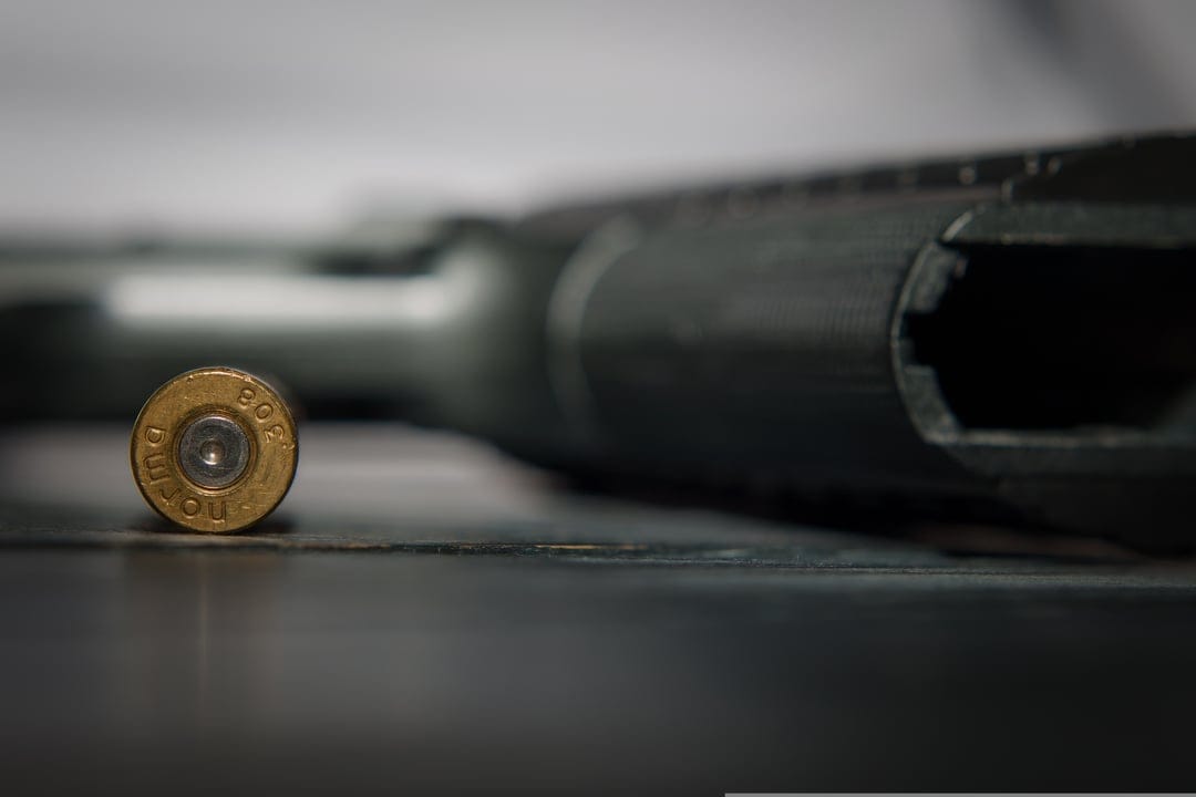 Brass bullet casing with caliber markings resting on a dark surface in front of a blurred tactical firearm.