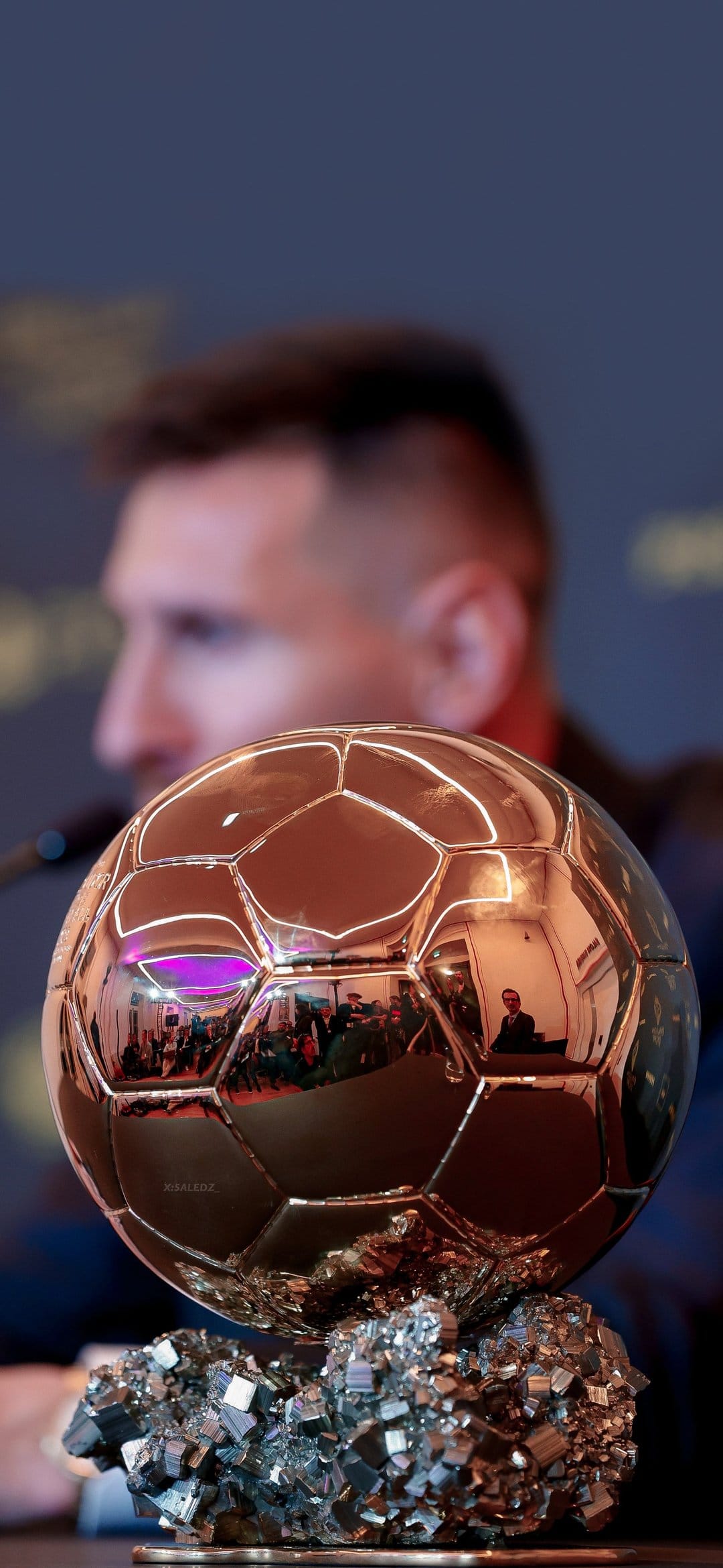 Lionel Messi blurred in the background behind a sharp golden football trophy with a crystalline base.