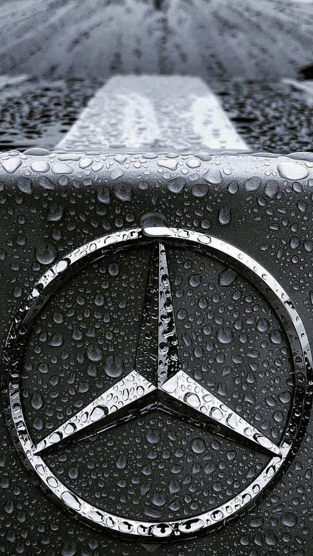 Mercedes-Benz star emblem covered in raindrops on a dark car hood with a blurred wet road in the background.