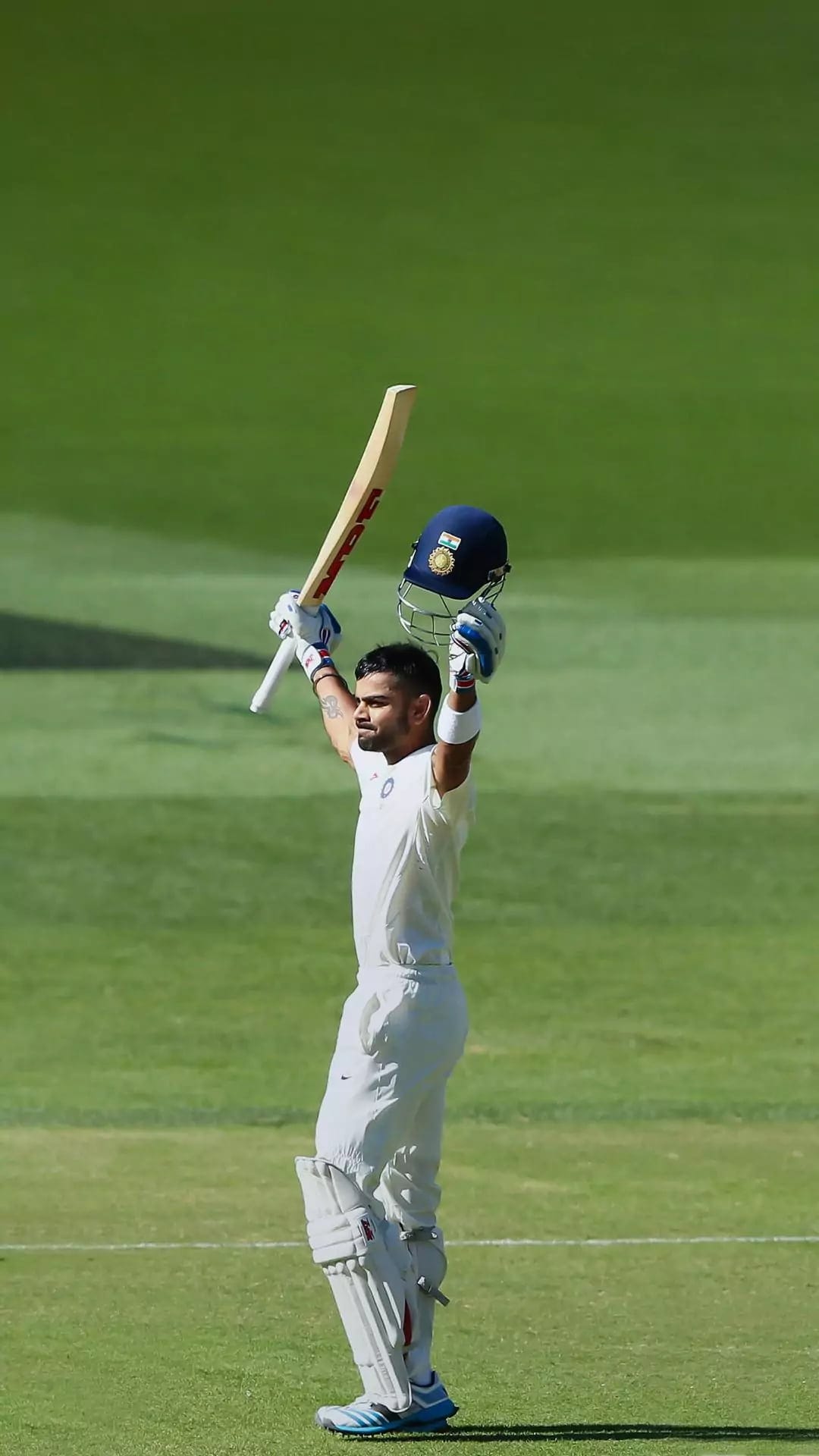 Virat Kohli raises his helmet and bat in celebration on a green field while wearing a white cricket uniform.