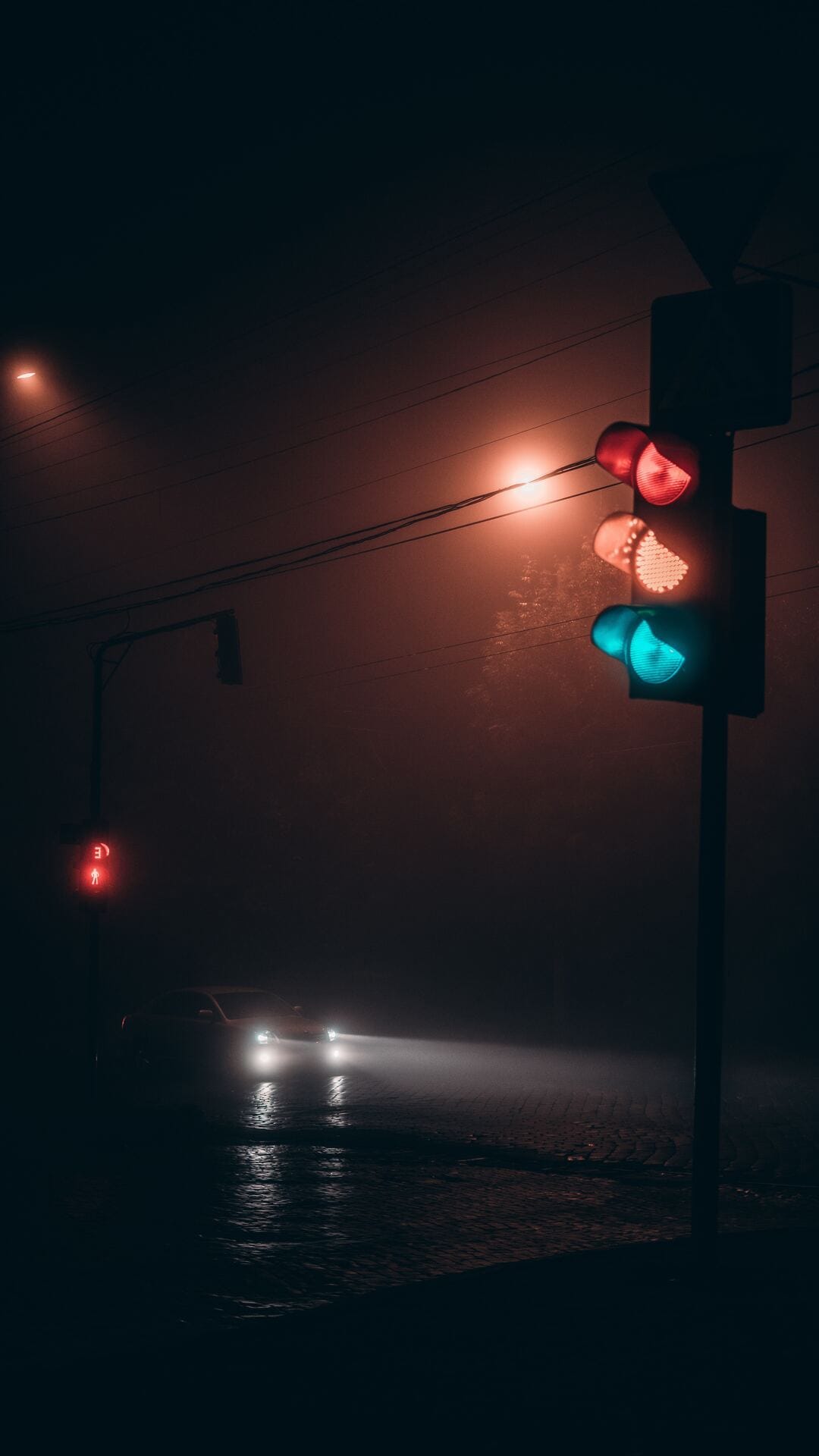 Traffic signals glow vividly through thick dark mist above a road with distant blurry headlights in the night.