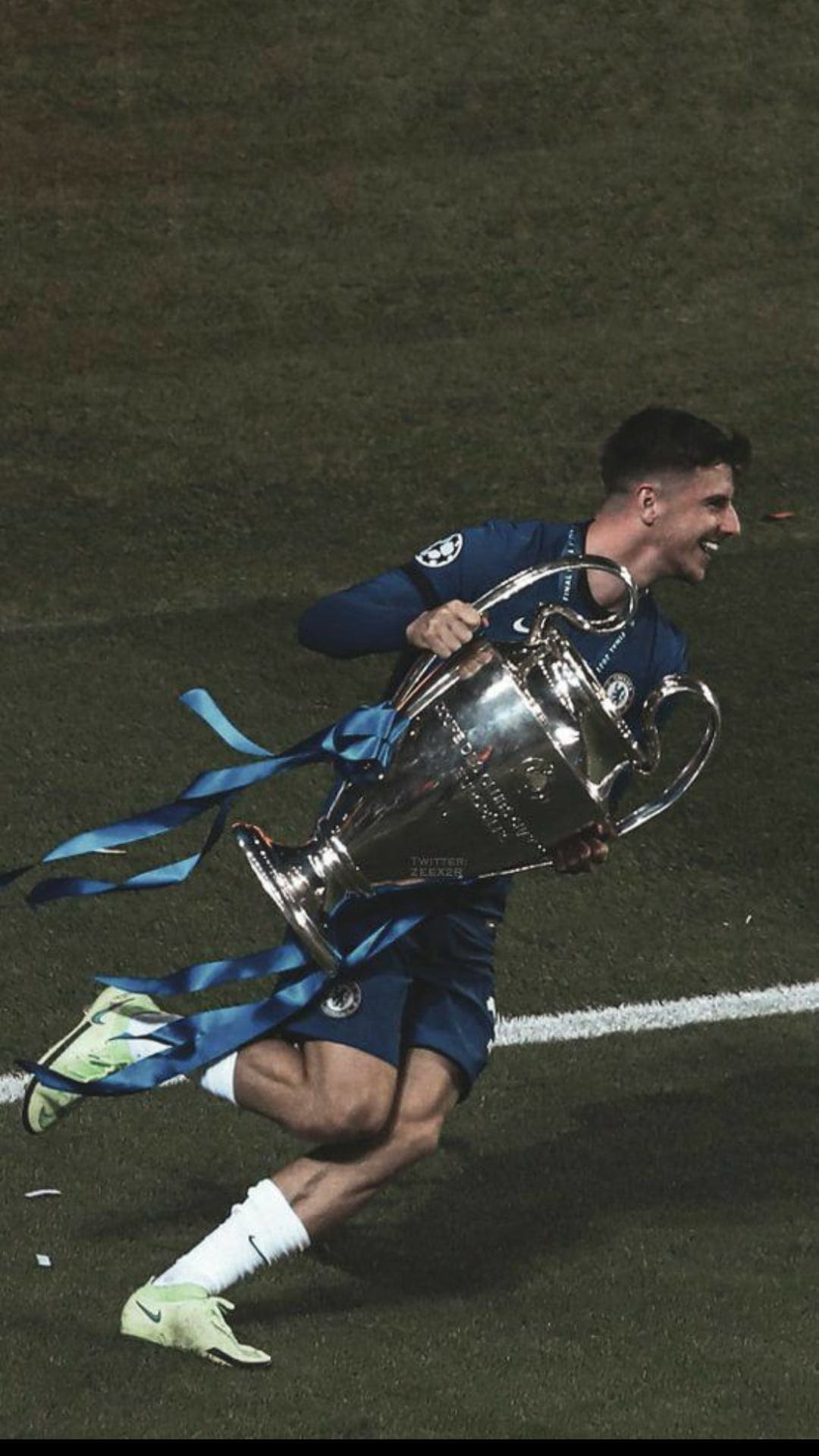 Mason Mount runs on a grass pitch while lifting the silver Champions League trophy with blue ribbons.