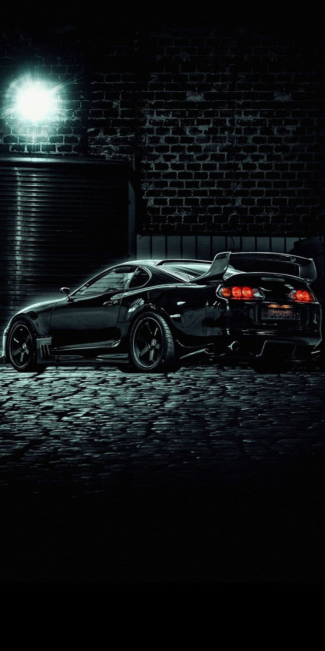 Black sports car parked on cobblestones under a bright spotlight against a weathered brick wall at night.