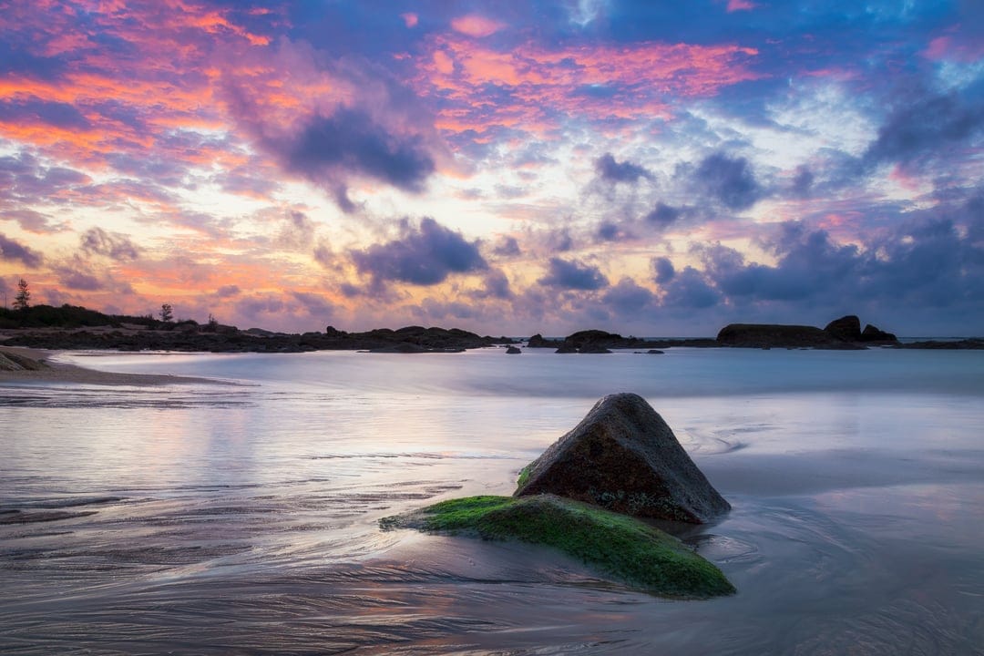 Mossy rocks sit on a wet sandy beach under a vibrant purple and pink sunset sky during a long exposure.