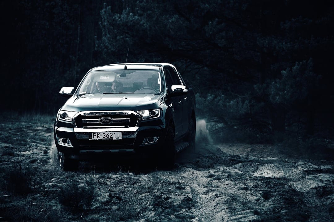 Ford Ranger truck with glowing headlights driving through a dusty dark forest on a rugged dirt path at night.