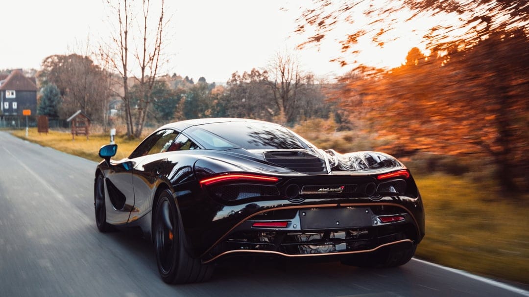 Rear view of a black McLaren 720S speeding on a rural road with autumn trees and golden sunset lighting.