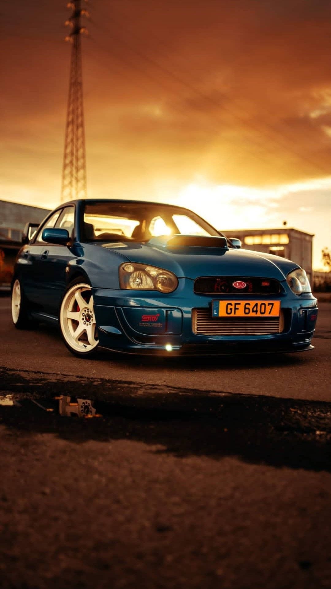 Blue Subaru WRX STI with white wheels parked on asphalt during a golden sunset with a warm orange sky.