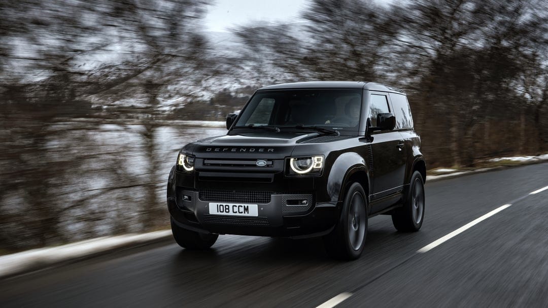 Black Land Rover Defender 90 driving on a wet road with motion blur and glowing circular LED headlights.