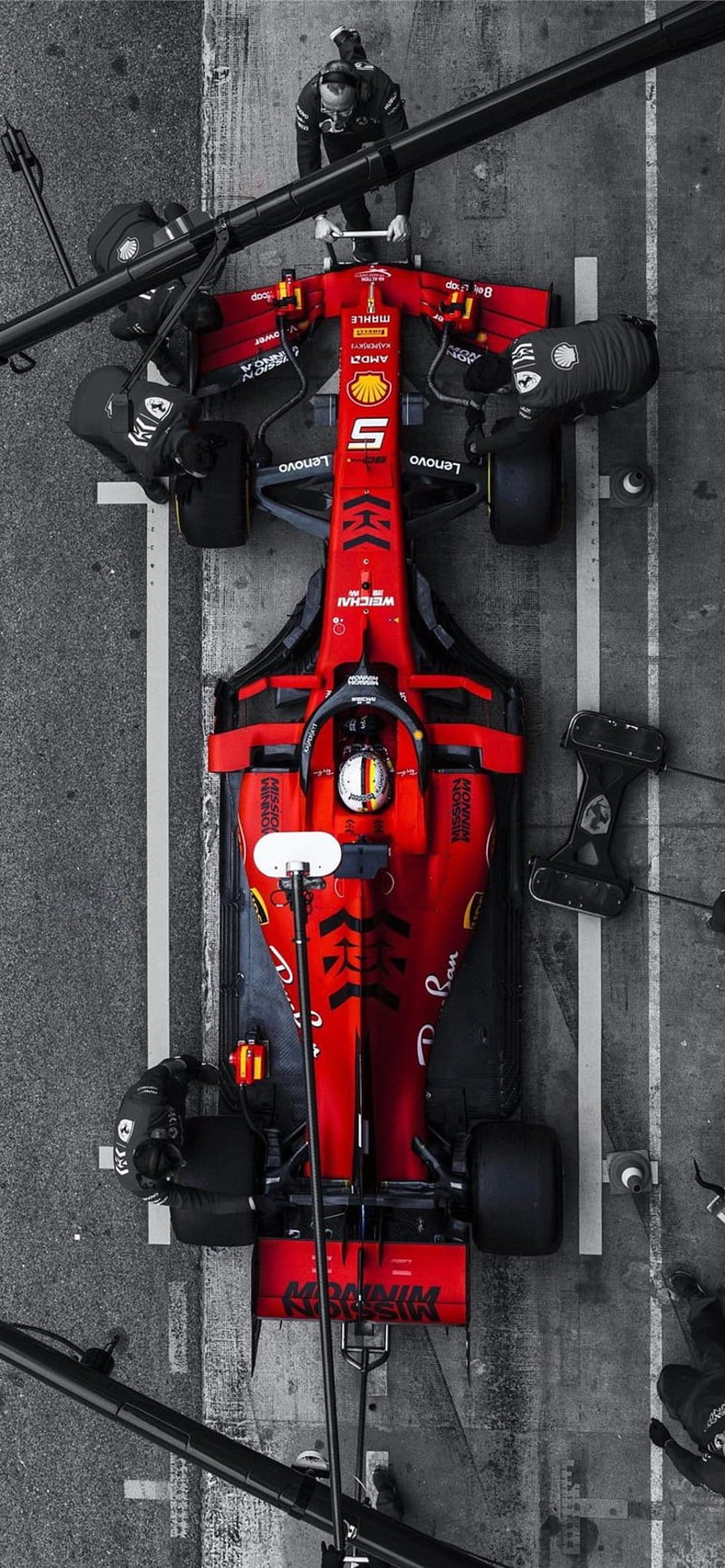 Red Ferrari Formula 1 car surrounded by pit crew mechanics in black suits on a grayscale pit lane asphalt.