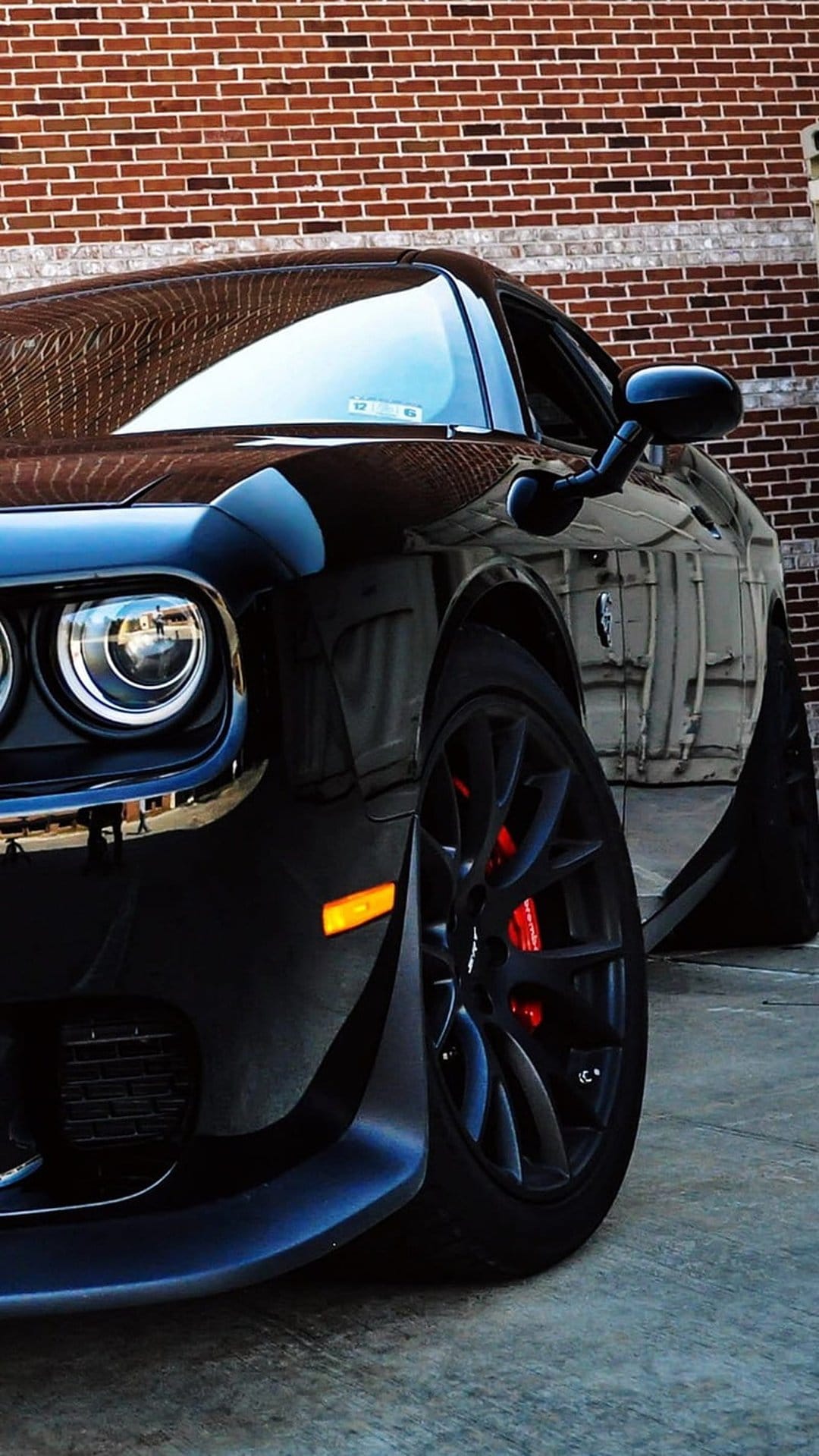 Black Dodge Challenger featuring round headlights and red brake calipers parked against a rustic brick wall.