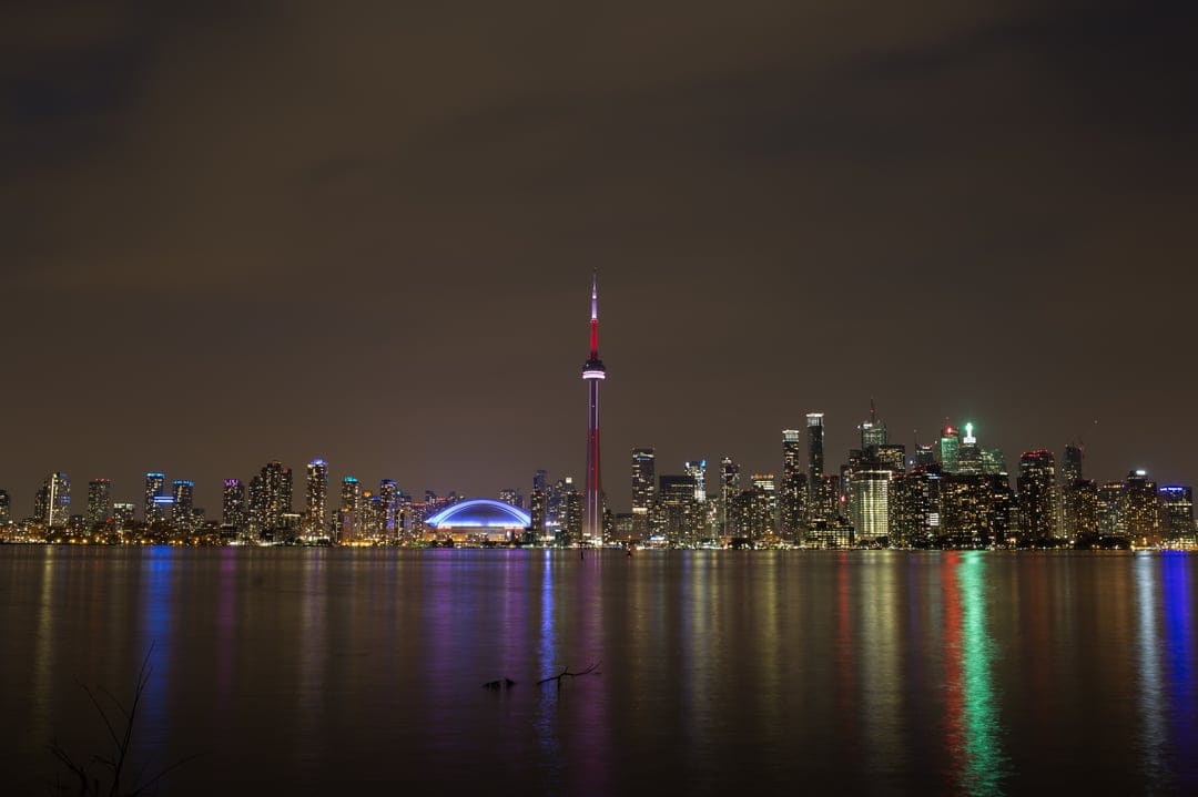 CN Tower and Toronto skyline reflected in dark lake water with glowing city lights under a clear night sky.