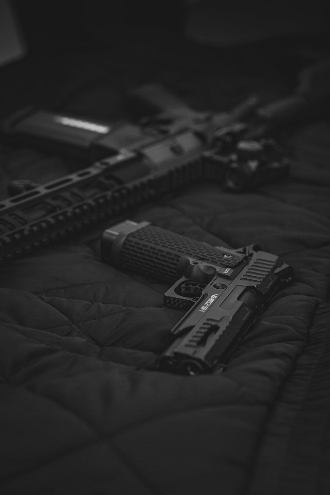 Tactical handgun with textured grips and a blurred rifle rest on a dark, moody grayscale metallic surface.