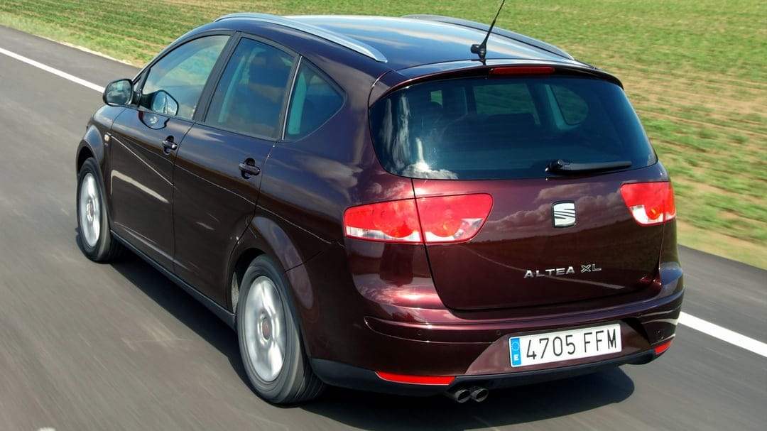 Rear view of a maroon SEAT Altea XL driving on a highway next to green fields in bright daylight.