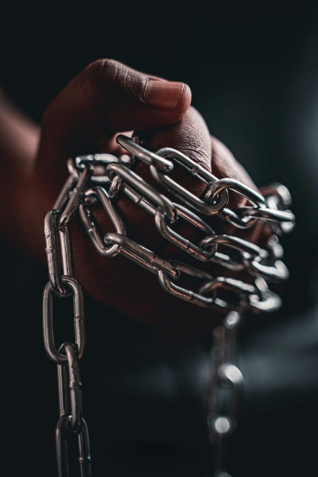 Man's hand tightly clenching a thick silver metal chain against a dark, out-of-focus industrial background.