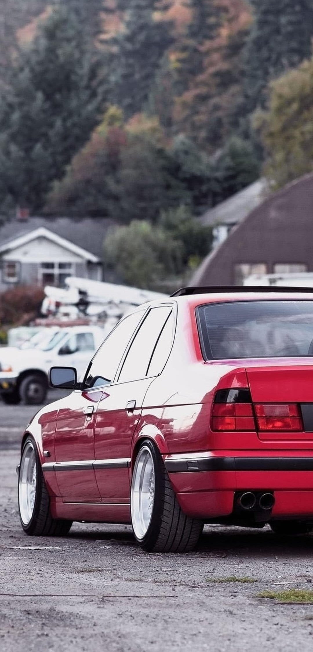 Red BMW sedan with polished chrome wheels parked on gravel against a suburban backdrop of green trees and houses.