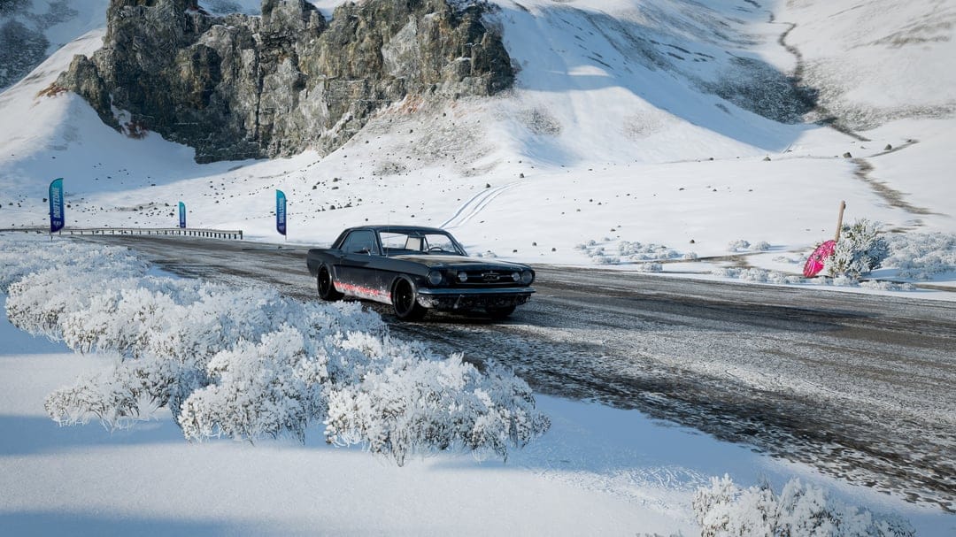 Black vintage Ford Mustang on a slushy mountain road surrounded by high snow-covered peaks and frozen bushes.