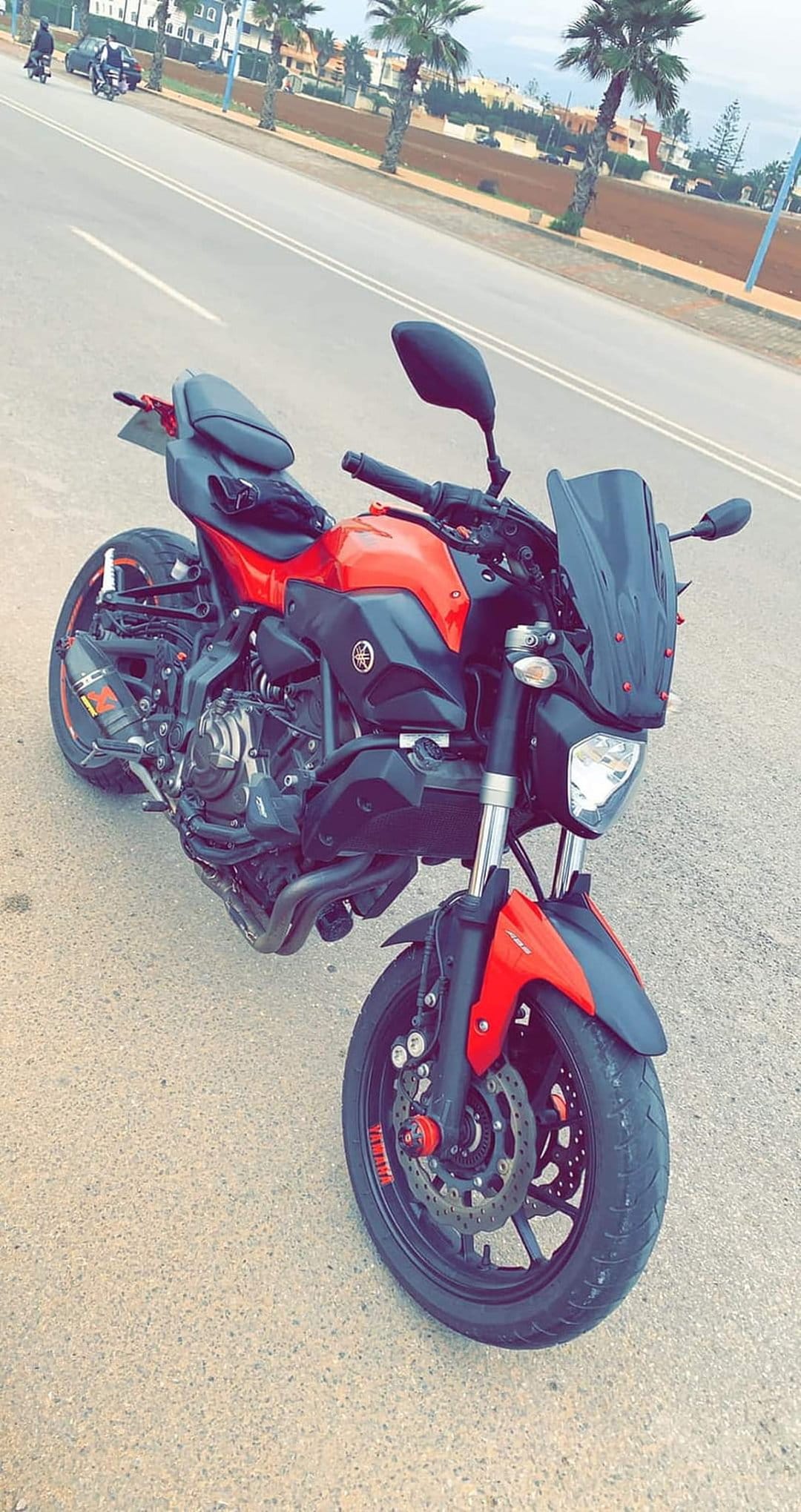 Red Yamaha motorcycle parked on an asphalt street with green palm trees and city buildings in the background.