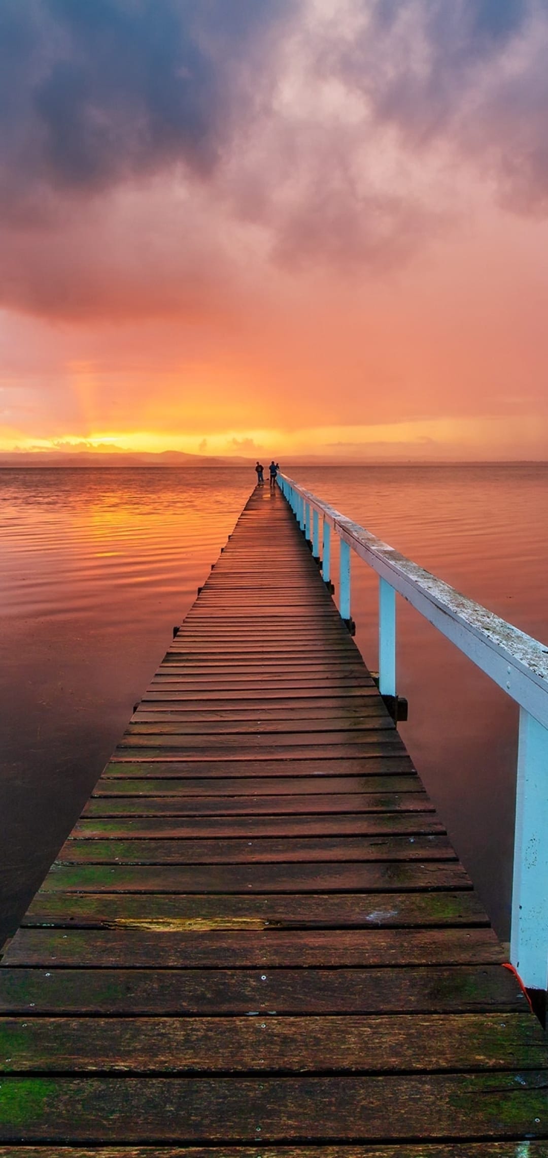 Wooden pier stretching toward a fiery orange sunset with silhouetted figures and calm water reflections.