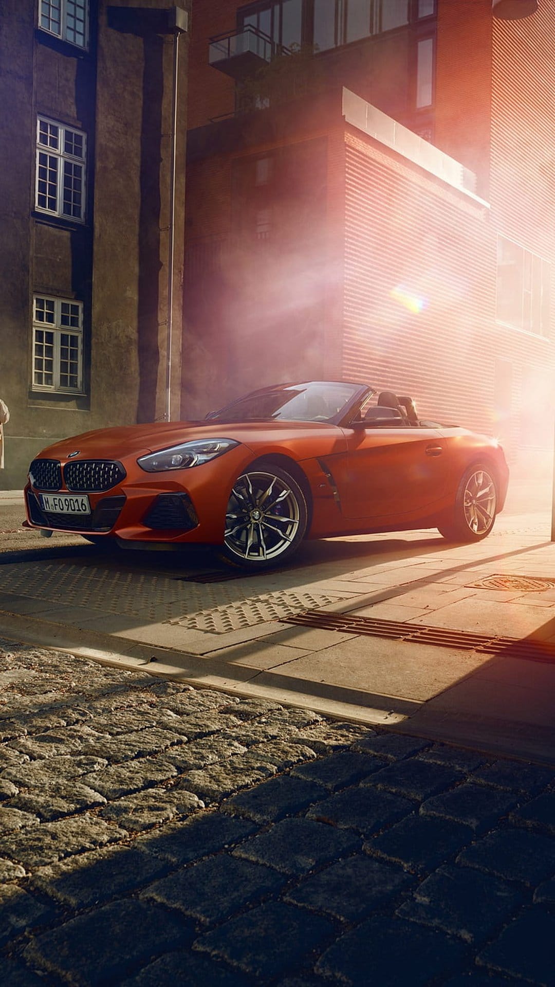 BMW Z4 Roadster in vibrant orange parked on a sunlit cobblestone street against a backdrop of modern buildings.