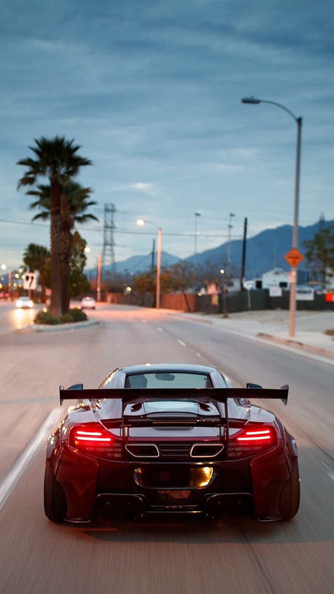 Red McLaren sports car with a large rear wing drives down a road lined with palm trees during a purple sunset.