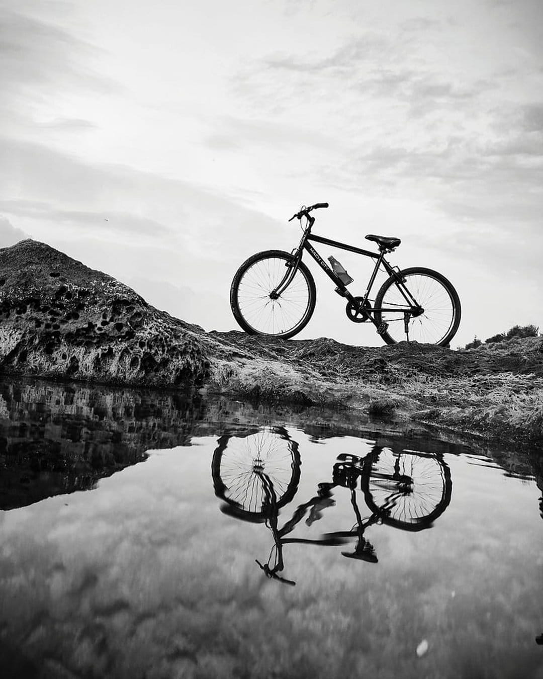 Vintage bicycle parked on a jagged rock formation reflecting in calm water under a dark, cloudy sky.