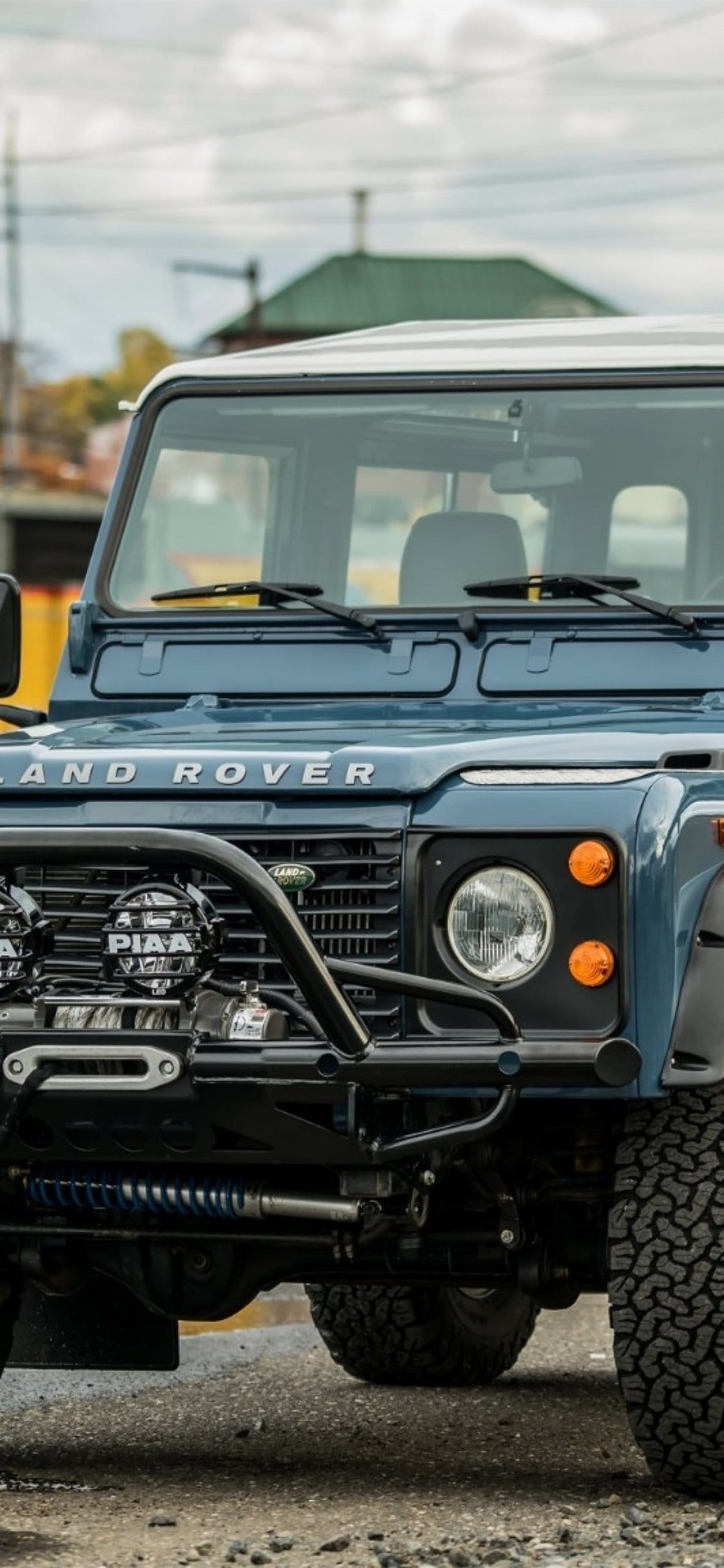 Front view of a blue Land Rover Defender with a winch, bull bar, and PIAA lights on gravel ground.