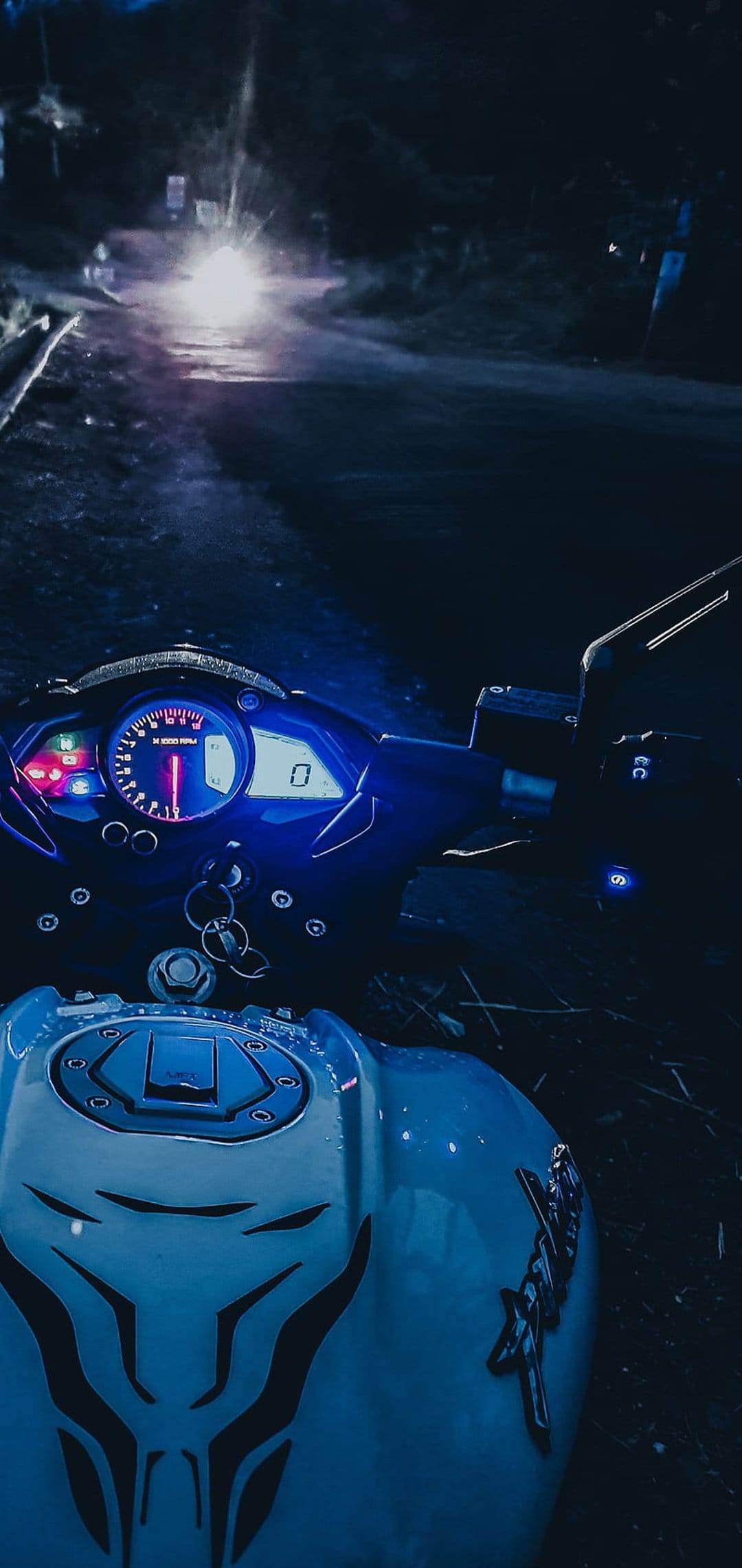 Motorcycle cockpit with glowing blue gauges and a tribal fuel tank view of a wet road at night from a rider POV.