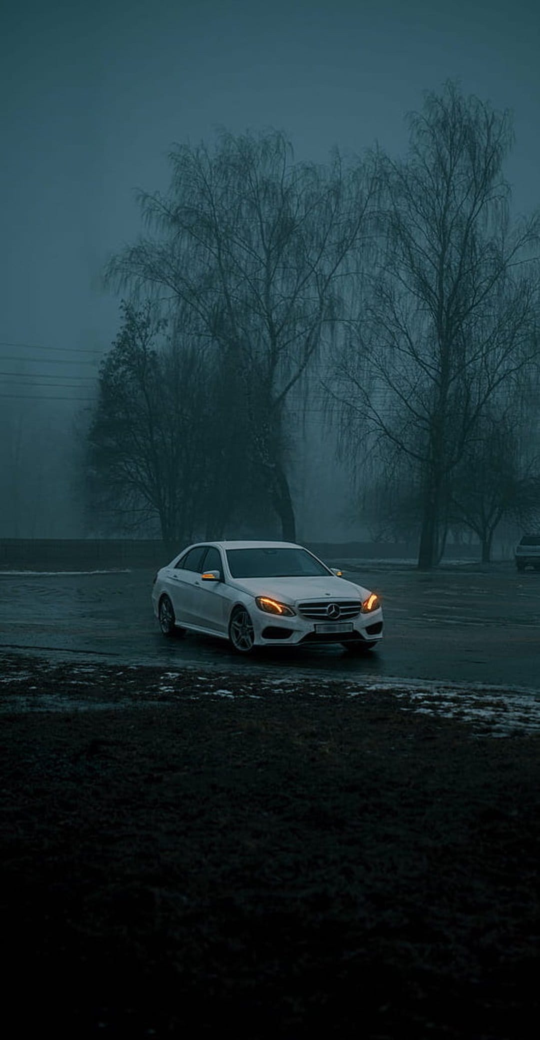 White Mercedes-Benz sedan with glowing headlights on a wet road surrounded by thick fog and bare winter trees.