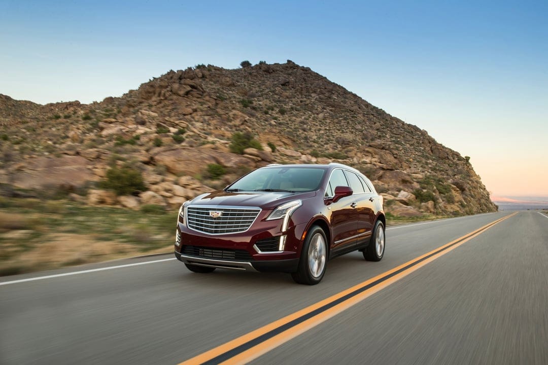 A dark red Cadillac SUV drives on an asphalt desert road past a large rocky hill under a clear sky.