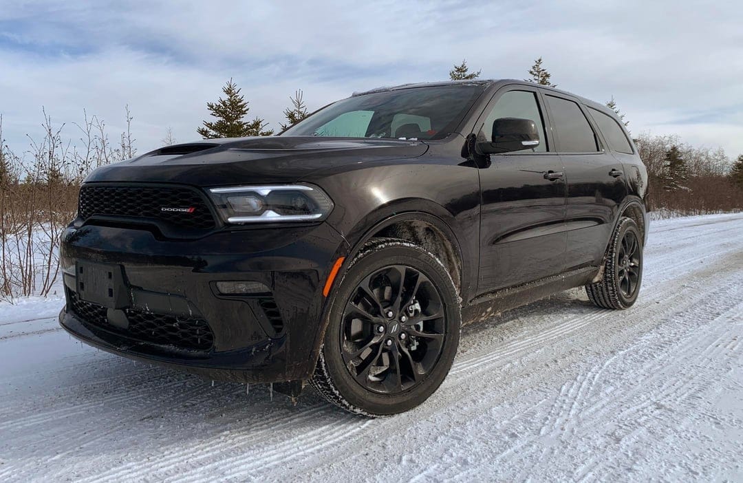Black Dodge Durango SUV with LED headlights parked on a snowy road with pine trees under an overcast sky.