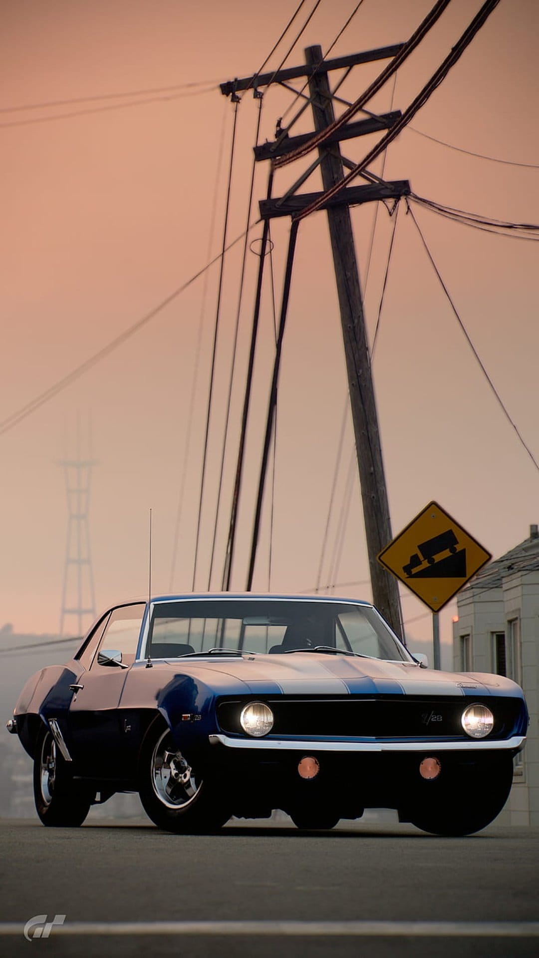 Blue vintage Chevrolet Camaro parked on a road with overhead power lines and a yellow steep grade sign at sunset.