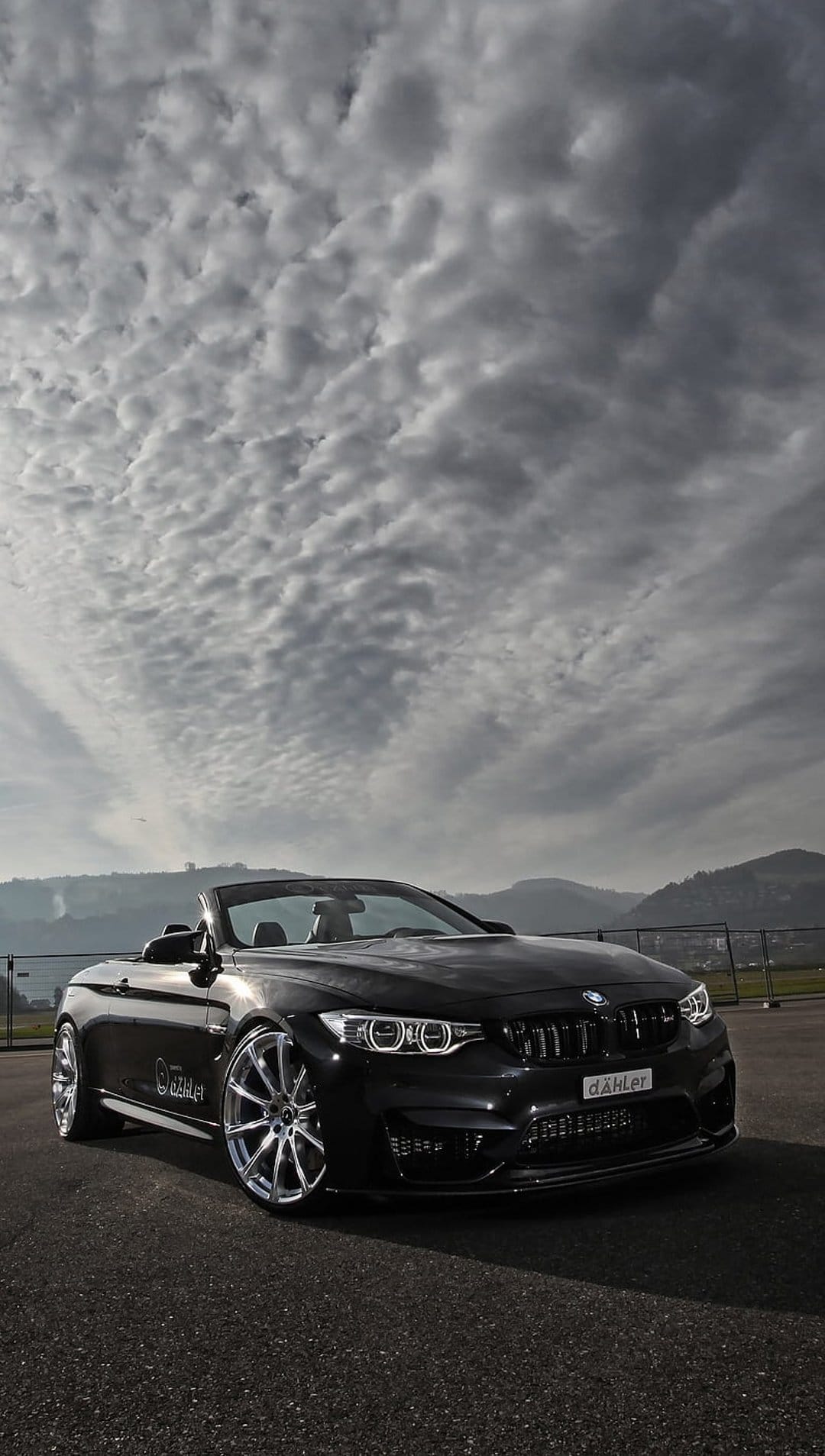 Black BMW M4 convertible seen from a low angle under a dark, cloudy sky with custom wheels and sleek bodywork.