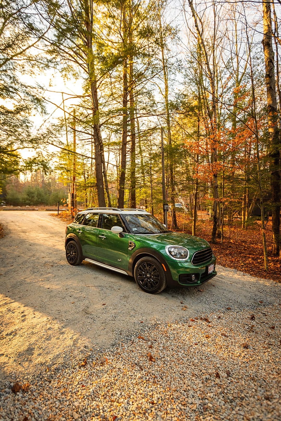 Green Mini Cooper Countryman parked on a gravel forest road with orange autumn leaves and golden hour sunlight.