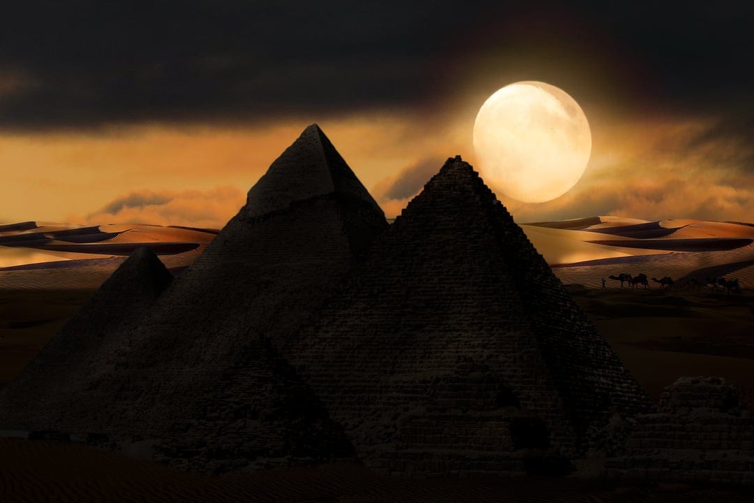 Great Pyramids silhouetted against a massive full moon in a starry night sky with camels on desert sand dunes.