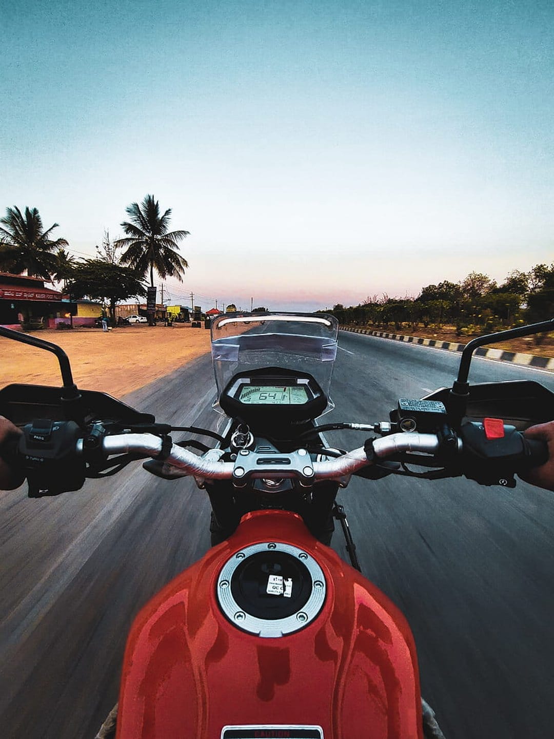 Red motorcycle handlebars on a highway at sunset with palm trees and a pink sky in a first-person perspective.