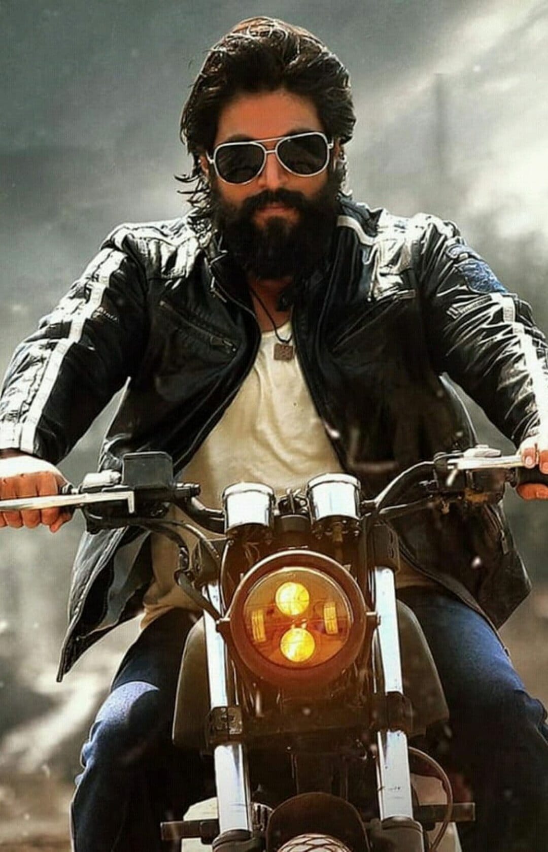 Bearded man wearing a black leather jacket and sunglasses rides a motorcycle with prominent headlights.