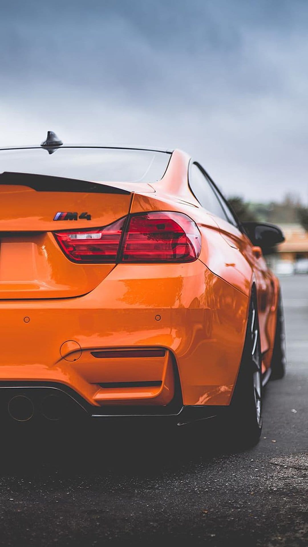 BMW M4 rear taillights and trunk badge on a bright orange sports car parked on dark asphalt under a gray sky.