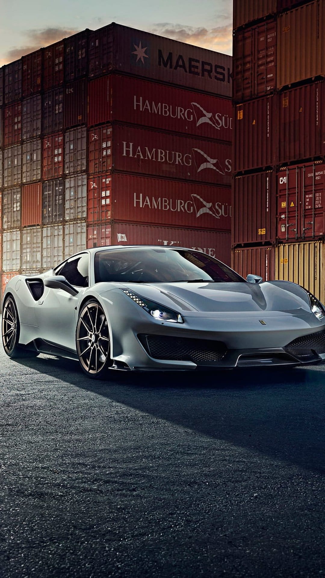 Ferrari 488 GTB in silver parked on asphalt before stacked red and blue shipping containers in a port setting.