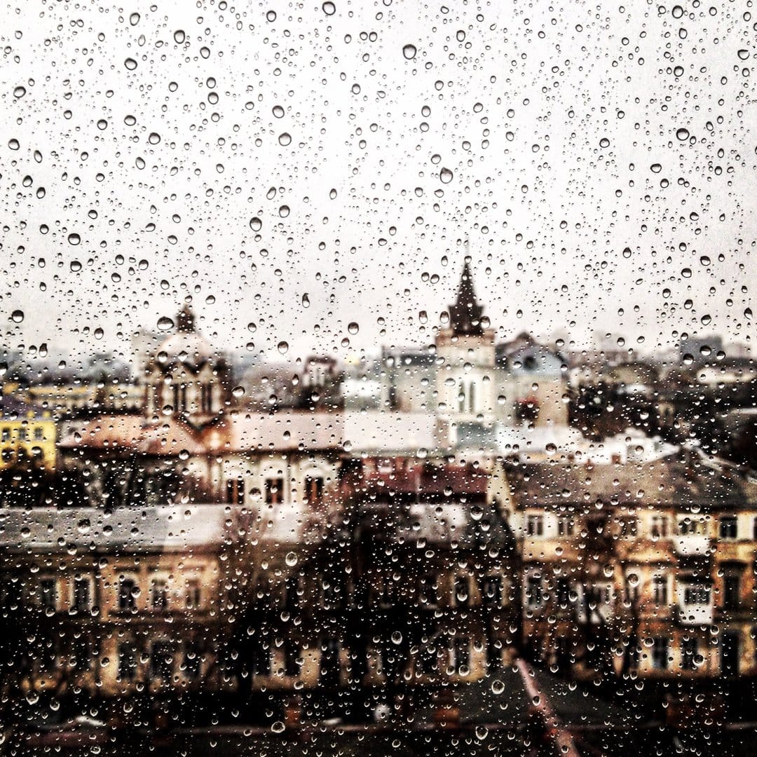 Raindrops on a glass windowpane frame a soft-focus view of historic European architecture and church spires.