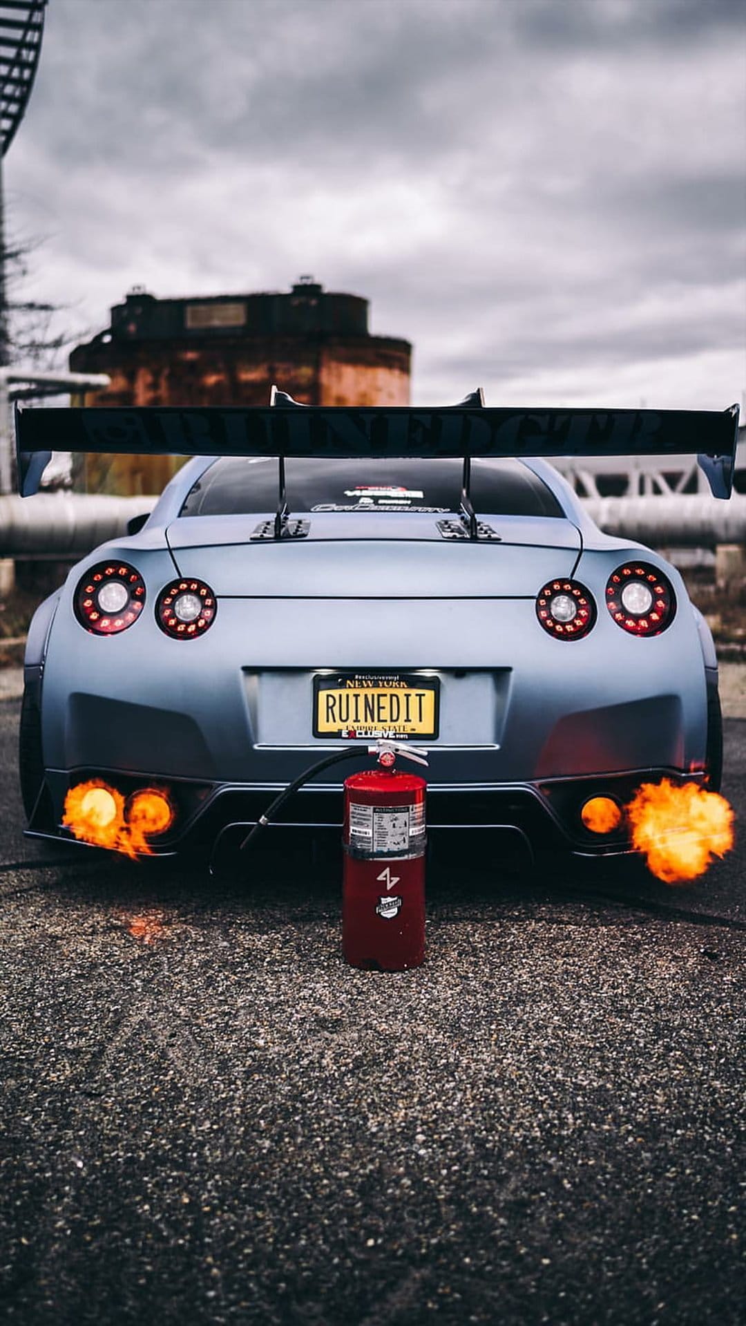 Nissan GT-R rear view featuring orange exhaust flames and a red fire extinguisher on asphalt during the night.