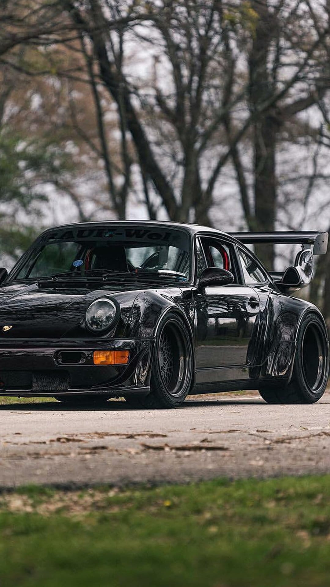 Porsche 911 widebody in black with a custom wing and dark wheels on an asphalt road near a forest background.