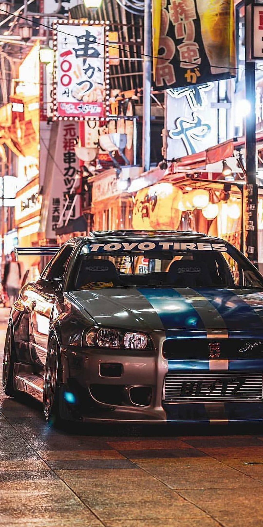 Nissan GT-R R34 with blue livery parked on wet cobblestones under glowing neon Japanese city signs at night.