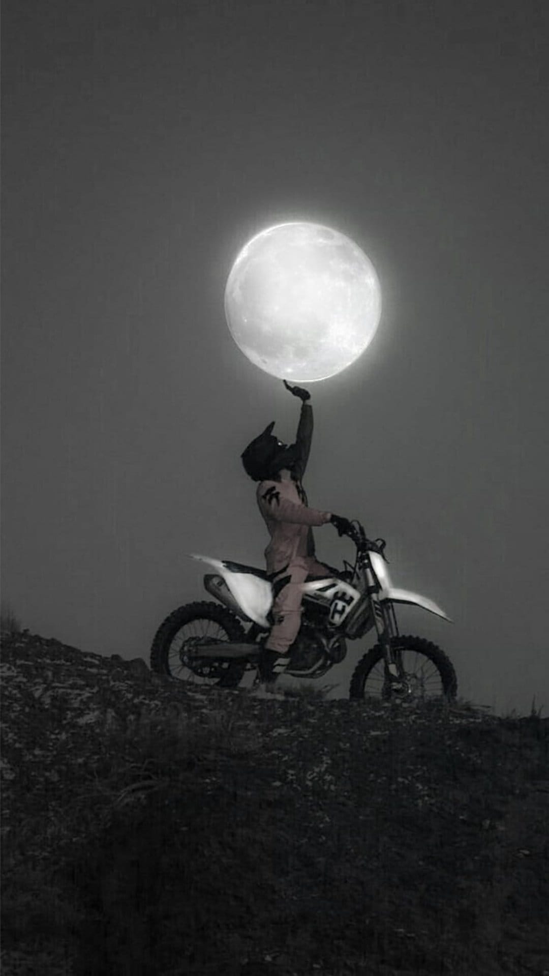 Motocross rider on a dirt bike reaches a hand toward a massive full moon under a dark, cloudy night sky.