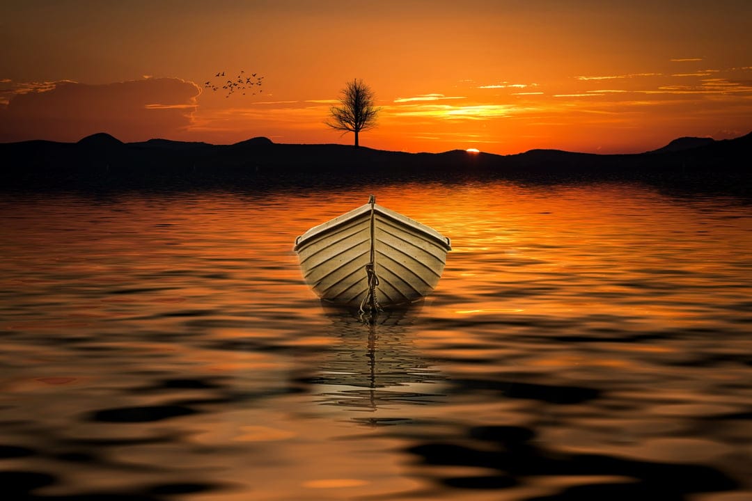 Small wooden rowboat on a still lake reflecting orange sunset clouds with distant hill silhouettes in the back.