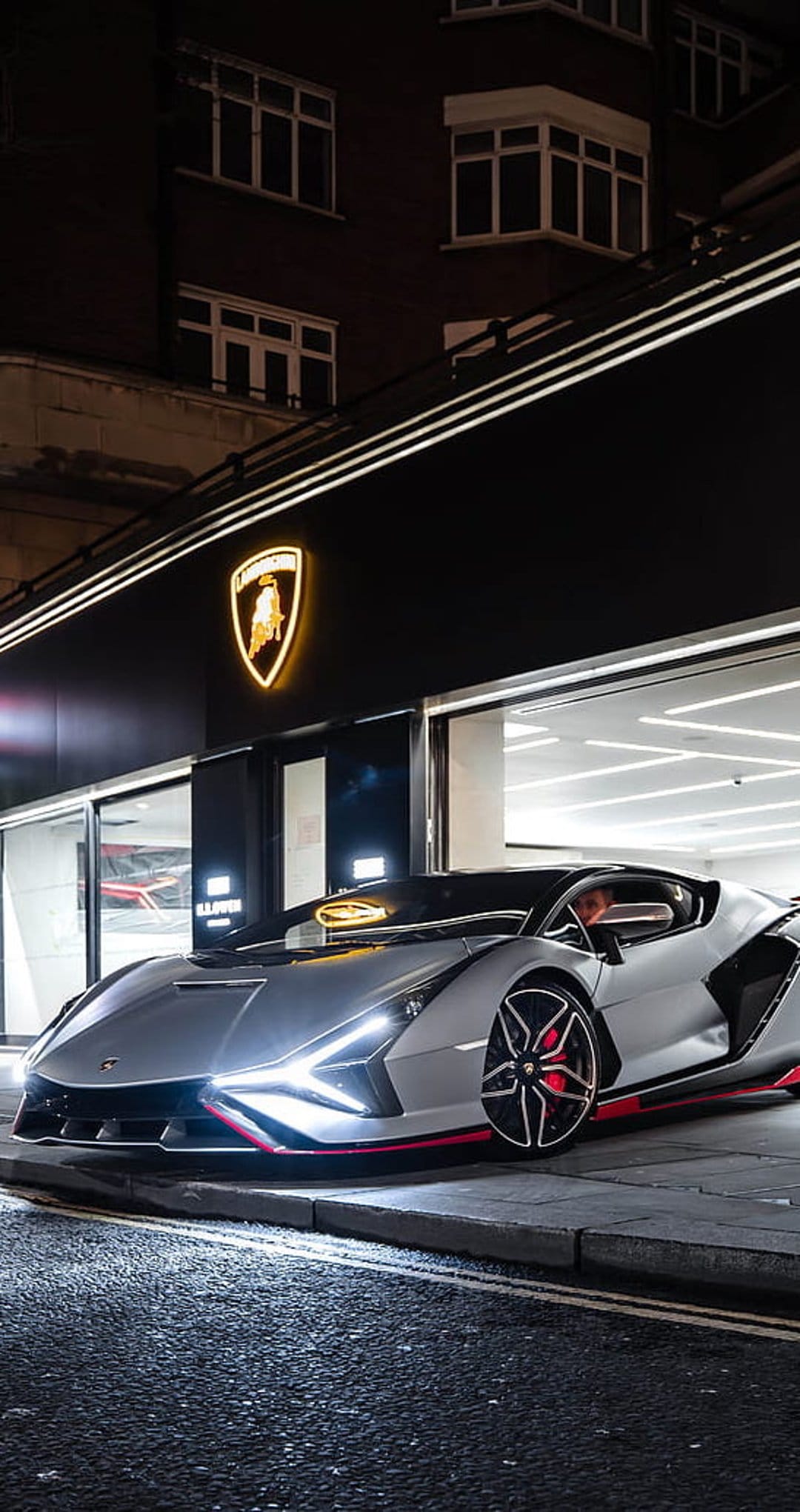Silver Lamborghini hypercar with glowing headlights parked outside a modern glass showroom with a lighted logo.