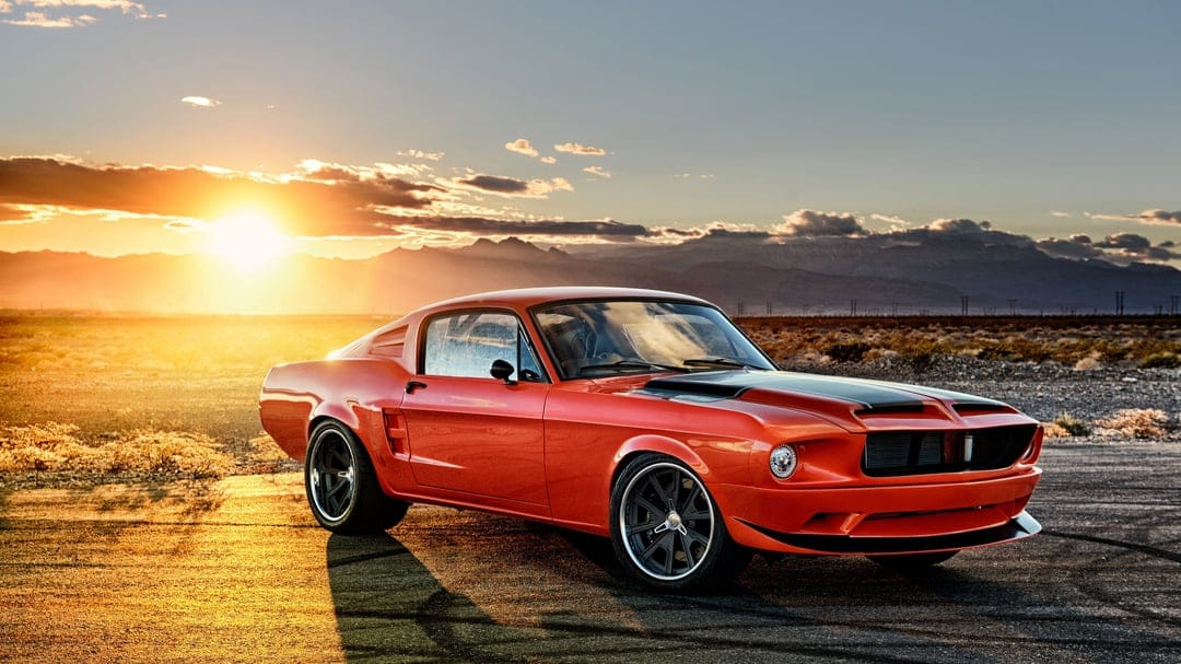 Orange classic muscle car with black wheels parked on a desert road during a bright golden sunset with mountains.