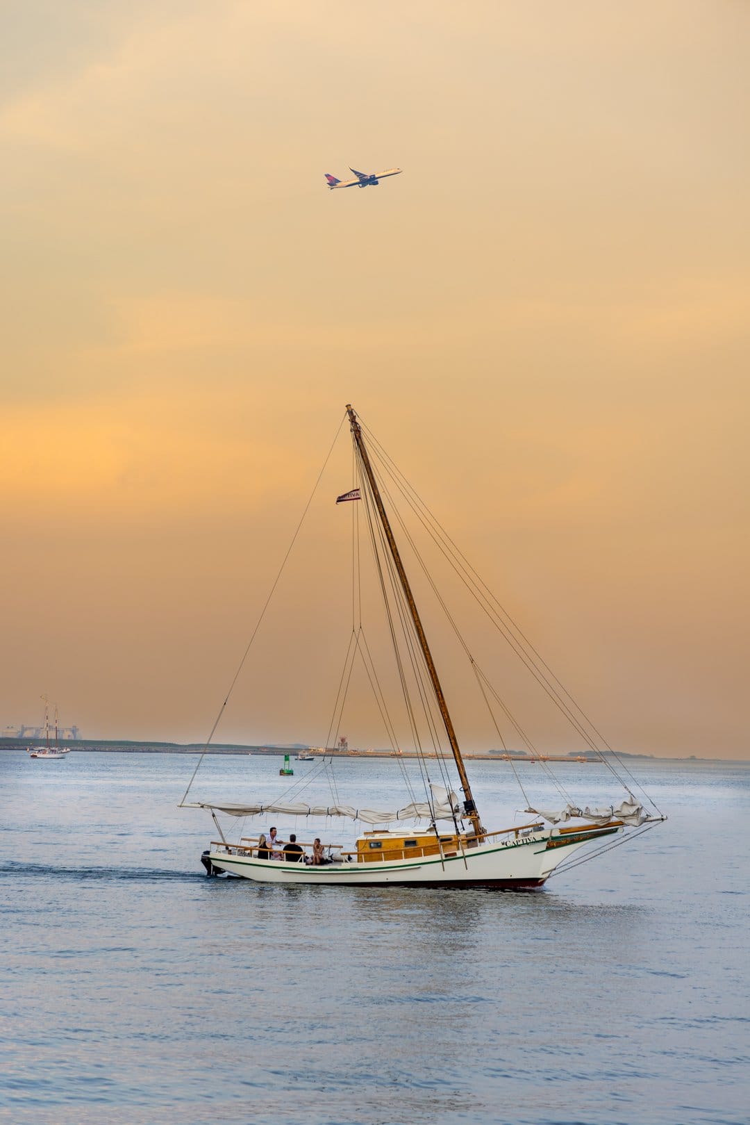 Sailboat drifting on calm water under a distant airplane during a vibrant orange and yellow sunset horizon.