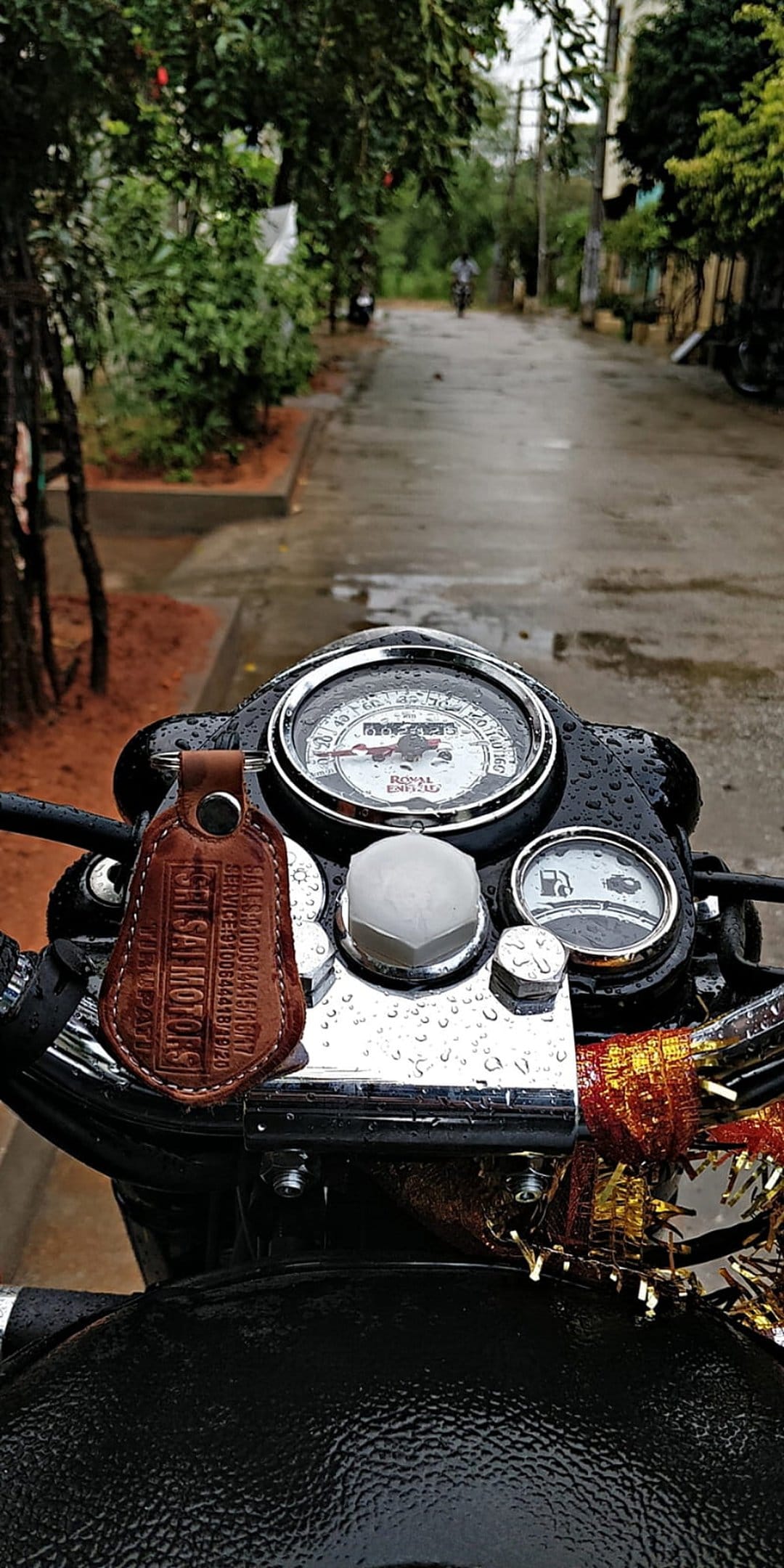 Motorcycle dashboard featuring a wet speedometer and fuel gauge before a blurred rainy road and green foliage.