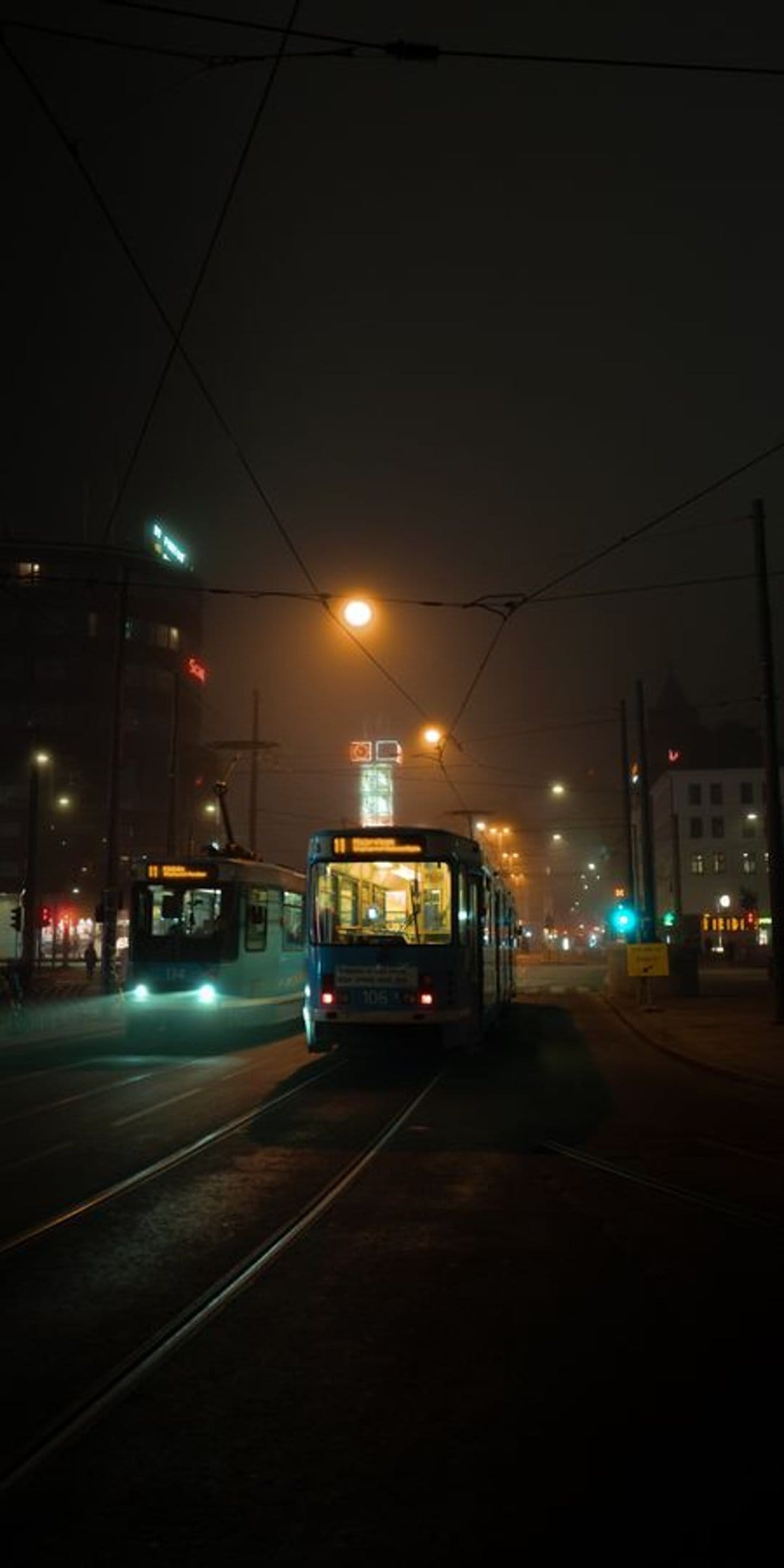 Modern trams with bright headlights travel down a misty city street at night over reflective wet asphalt.