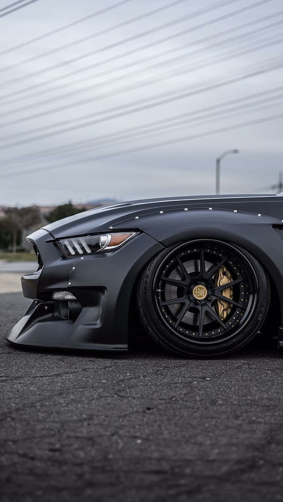 Matte black Ford Mustang with widebody fender flares, gold brake calipers, and a lowered stance on asphalt.